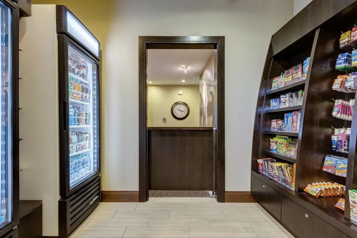 A refrigerated beverage display and snack shelf occupy one wall, with a doorway leading to another space and a wall clock above. The layout suggests convenient access to drinks and snacks, reinforcing the idea of flexible, in-room or on-the-go dining.