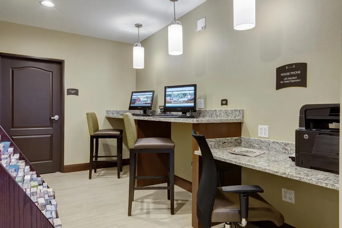 A functional and welcoming business center outfitted with computers, a granite countertop, bar stools, a printer, and a house phone. The space is arranged for easy access to business services and casual social interaction.
