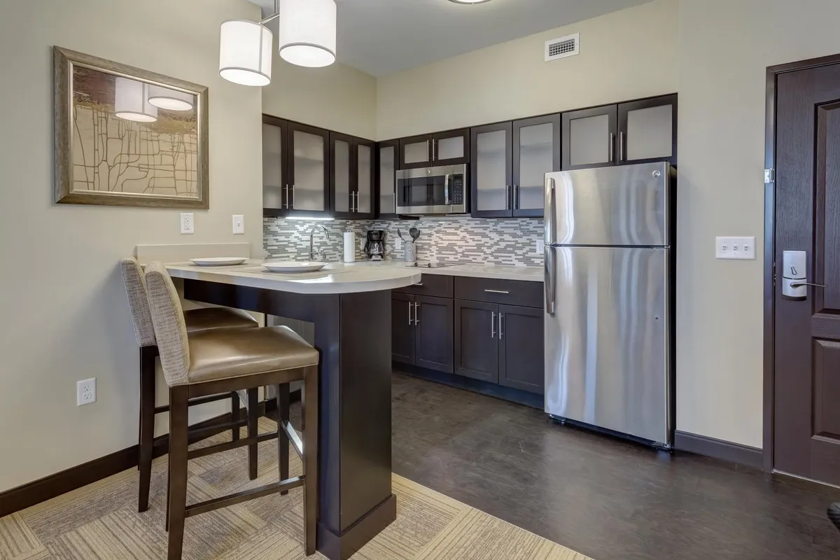 A modern kitchen area is highlighted by a central island, stainless steel appliances, contemporary light fixtures, and a textured backsplash. The space is open and inviting, suggesting opportunities for casual dining, sharing meals, or simply enjoying the comforts of a well-appointed suite kitchen.
