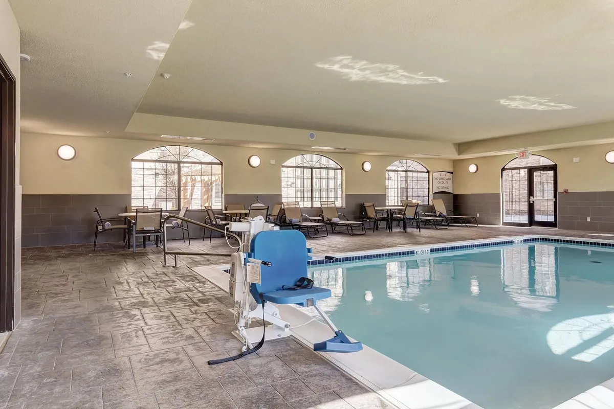A bright indoor swimming pool space with large windows letting in natural light, featuring an accessible pool lift, lounge chairs, and a calm, welcoming atmosphere. The scene highlights relaxation and recreation options for guests.