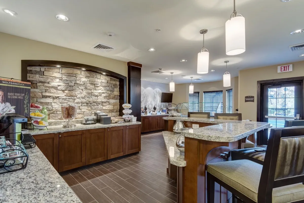 This image features a spacious interior with a granite countertop and a textured stone wall, softly illuminated by modern lighting fixtures. The setting is warm and welcoming, suggesting a communal dining or breakfast area where guests can enjoy meals together.