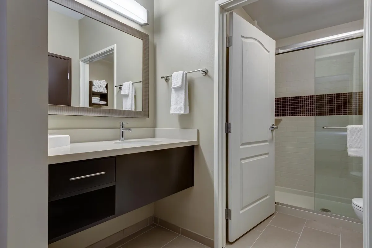 A calming bathroom scene features a vanity with a large mirror, modern sink, and a shower area. The space is bright and spa-like, suggesting a refreshing morning routine or a relaxing end to a busy day.