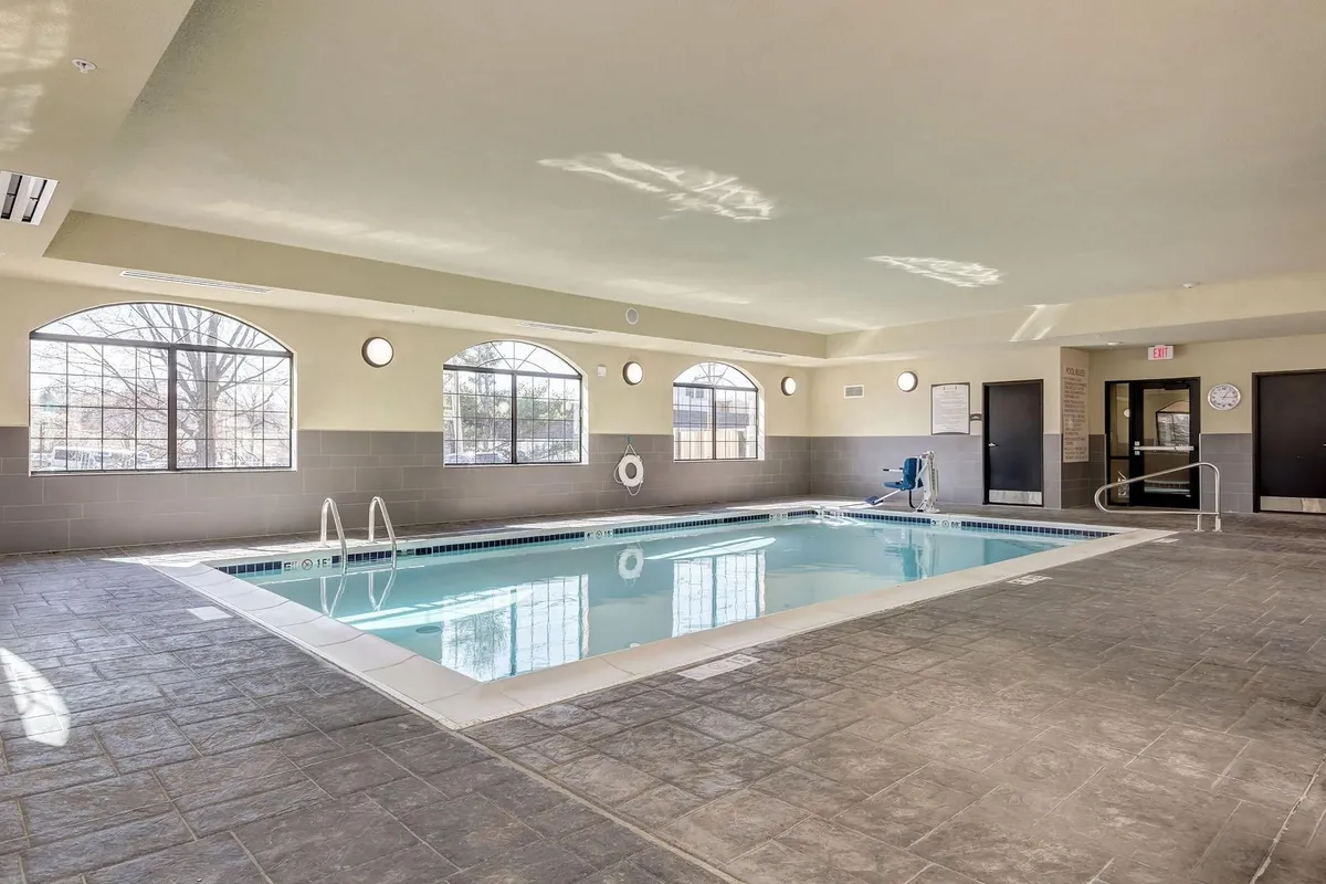 A bright, tranquil indoor pool space with large windows letting in natural light. The pool is surrounded by poolside seating and accessibility equipment, making it welcoming to all guests. The water shimmers with sunlight, and there’s an inviting atmosphere for relaxation or laps.