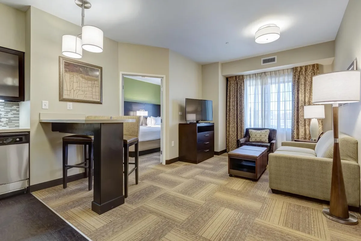 The image shows a cozy, well-lit condo interior featuring a stylish bar table with stools perfect for sharing meals or drinks, a comfortable sofa facing a large television for relaxing evenings, and a window with flowing curtains that lets in natural light—creating an inviting, home-like atmosphere for guests.
