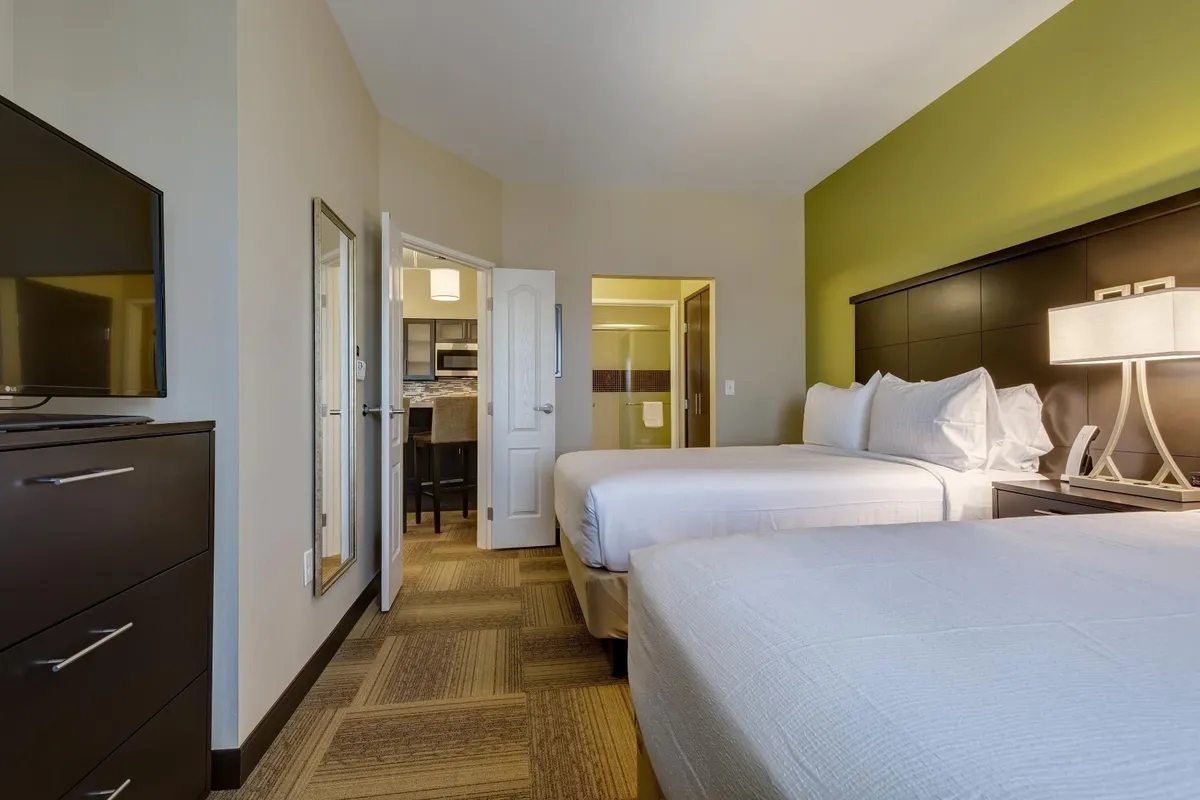 This image shows a spacious bedroom featuring two queen beds adorned in crisp white linens. A nightstand with a lamp sits between the beds for convenience, while a flatscreen TV is positioned for easy viewing from the beds. The room includes a mirror and a doorway leading to a kitchenette, providing a glimpse of the suite's added functionality.