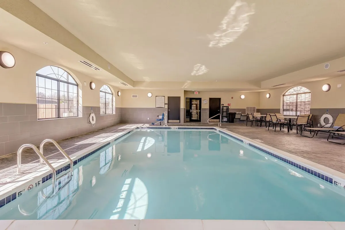 An inviting indoor swimming pool area featuring sparkling blue water, comfortable lounge chairs lining the sides, and large windows that let in natural sunlight. Poolside stairs offer easy access, and the tranquil atmosphere is ideal for relaxation and unwinding.